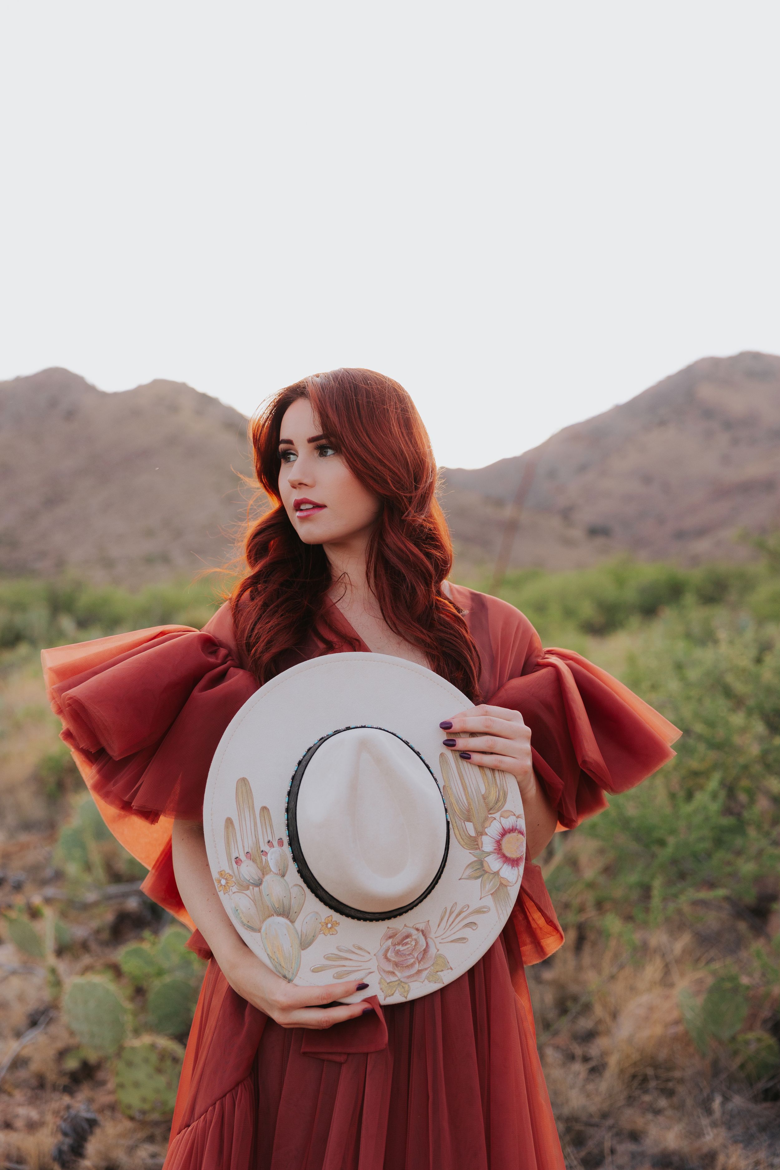 Artistic desert portrait with decorative wide-brimmed hat and burgundy dress.