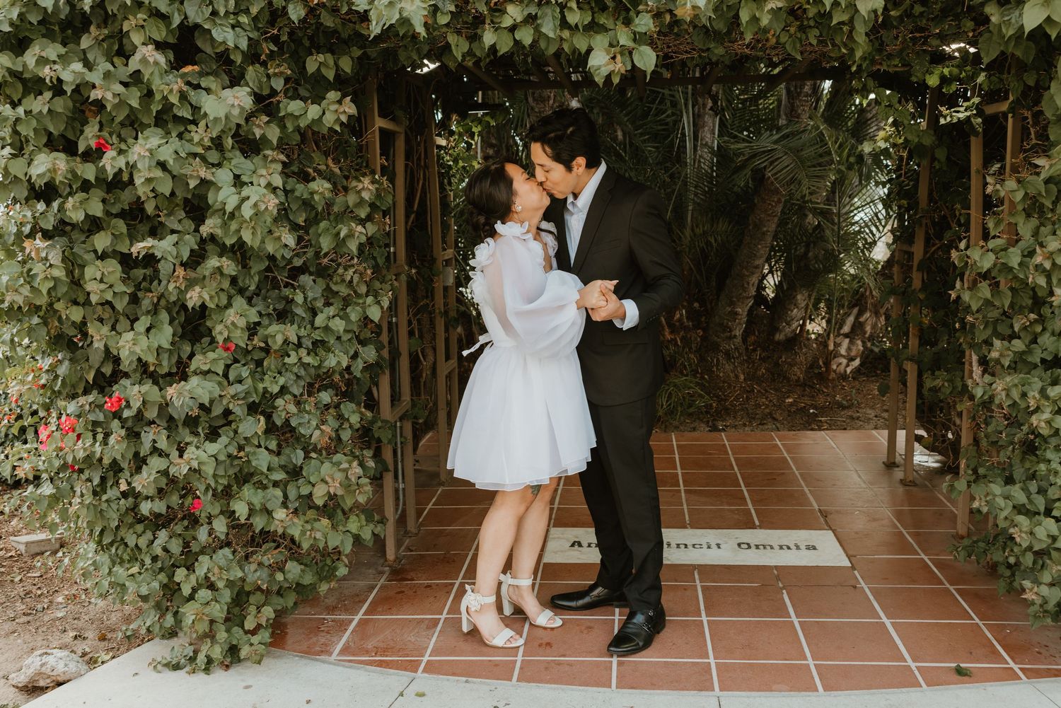 A romantic couple in formal attire embraces each other on a tiled patio surrounded by lush green foliage at the San Diego Courthouse.