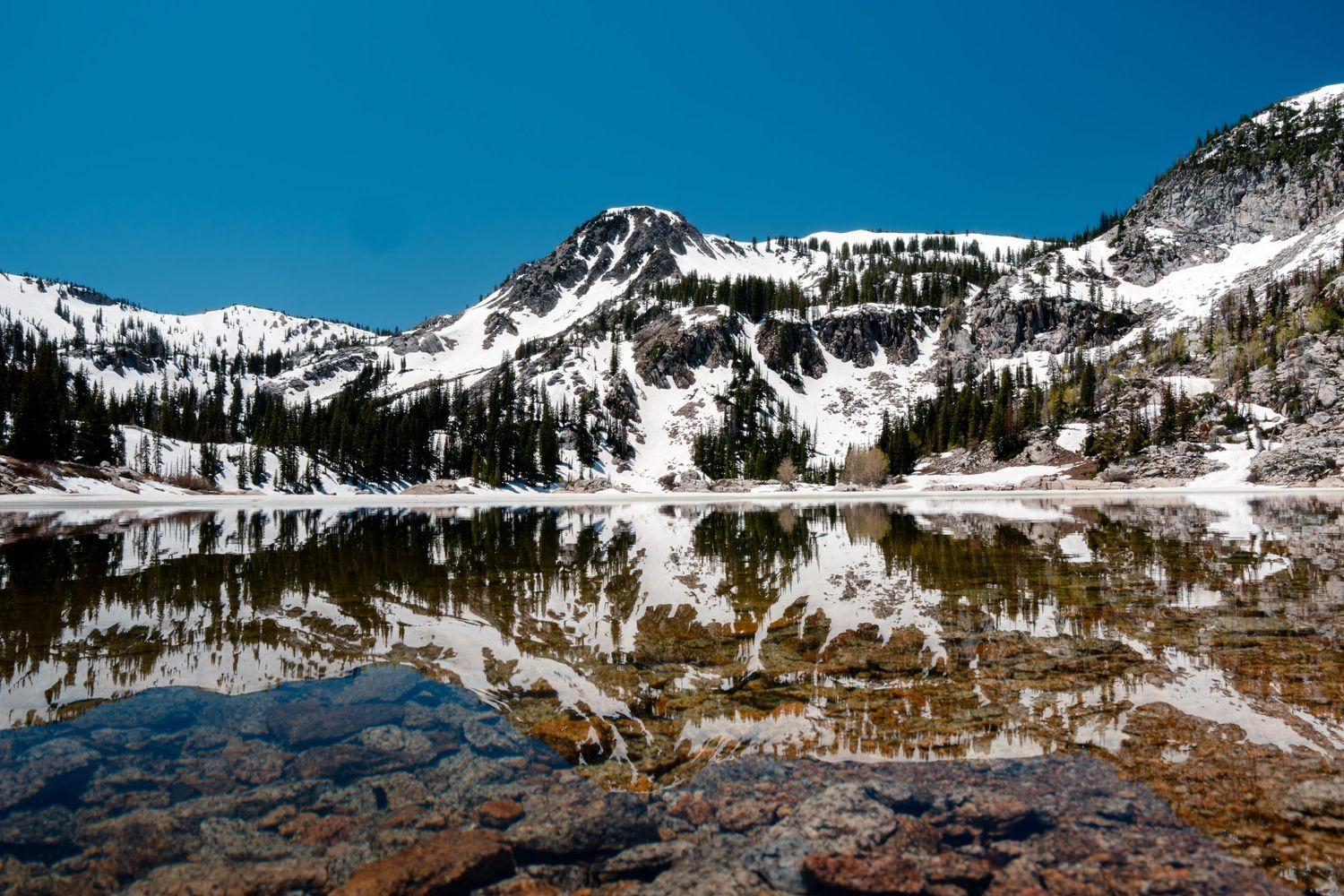 Snow-capped mountains reflecting in crystal clear alpine lake under bright blue sky.