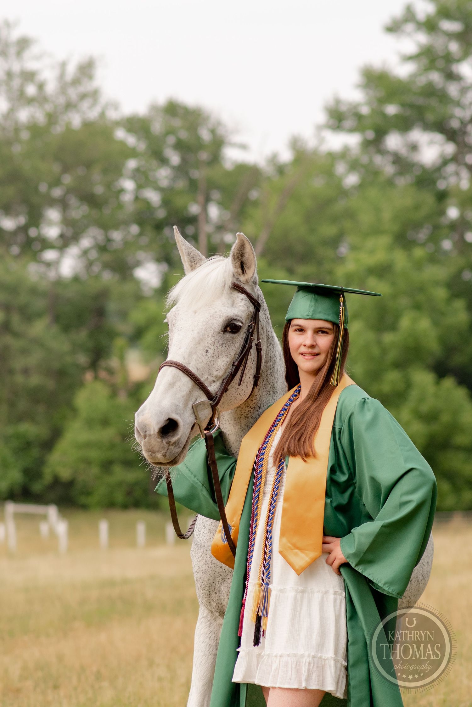 Graduate wearing green cap and gown poses with white horse against leafy background.