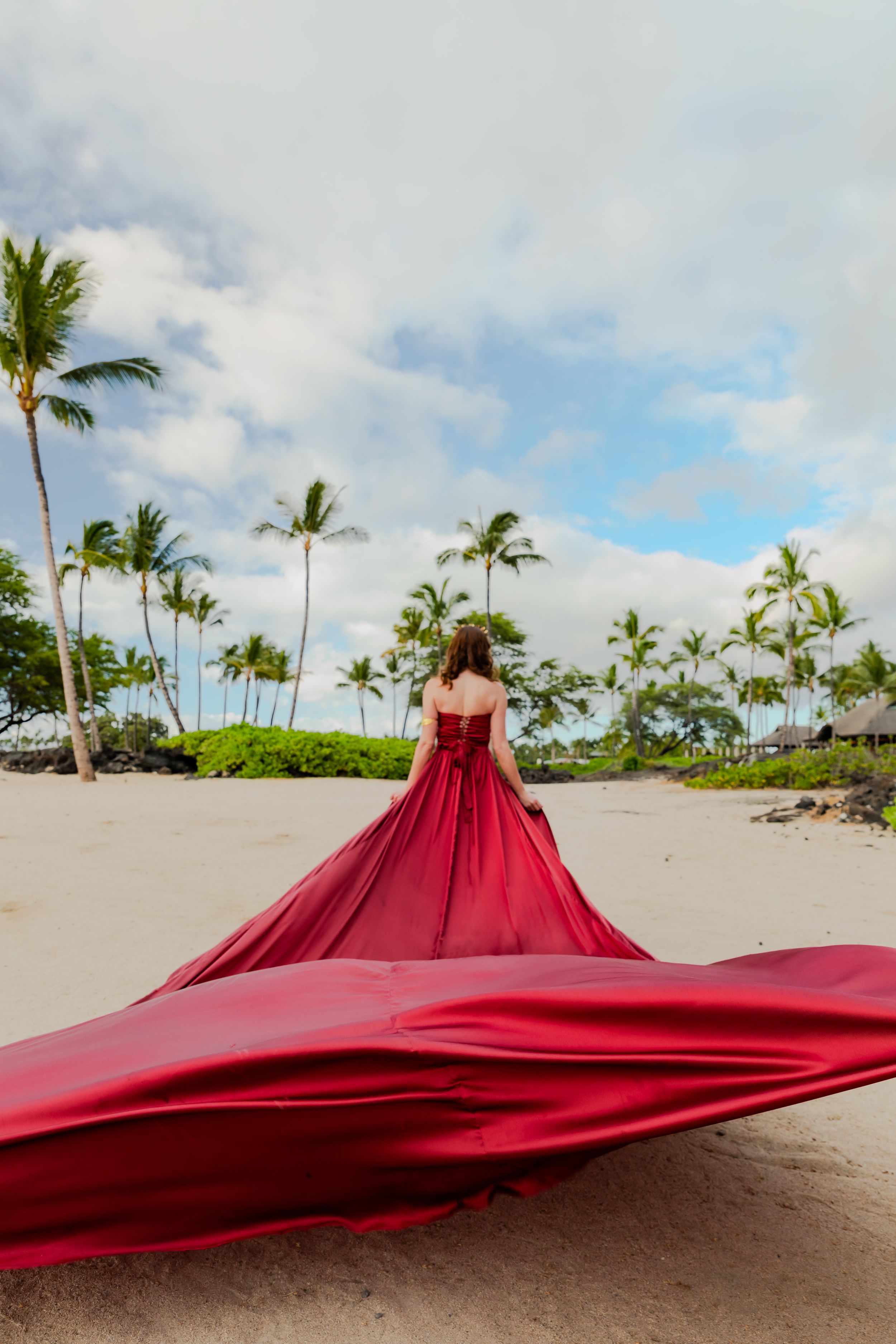 A dramatic red dress flows in the wind on a Hawaiian beach with swaying palm trees.