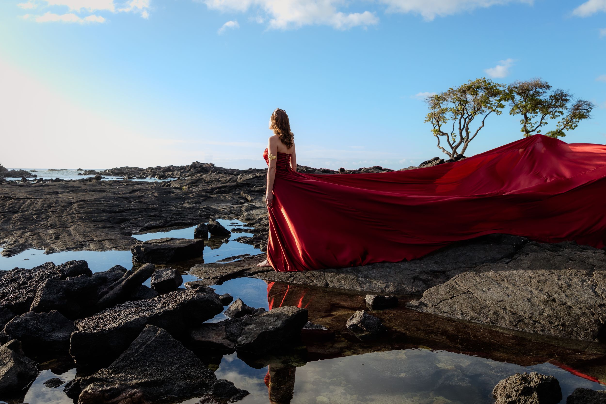 A red dress flows dramatically against volcanic rocks and ocean at sunset.