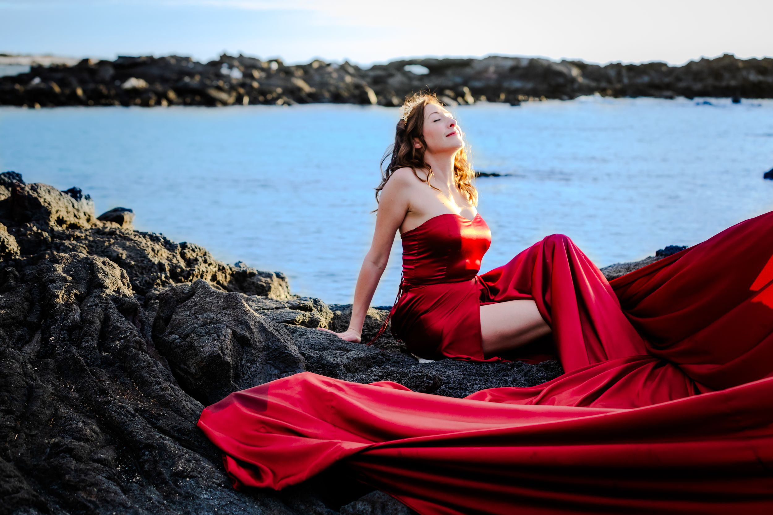 A red gown contrasts against black volcanic rocks beside blue ocean waters.