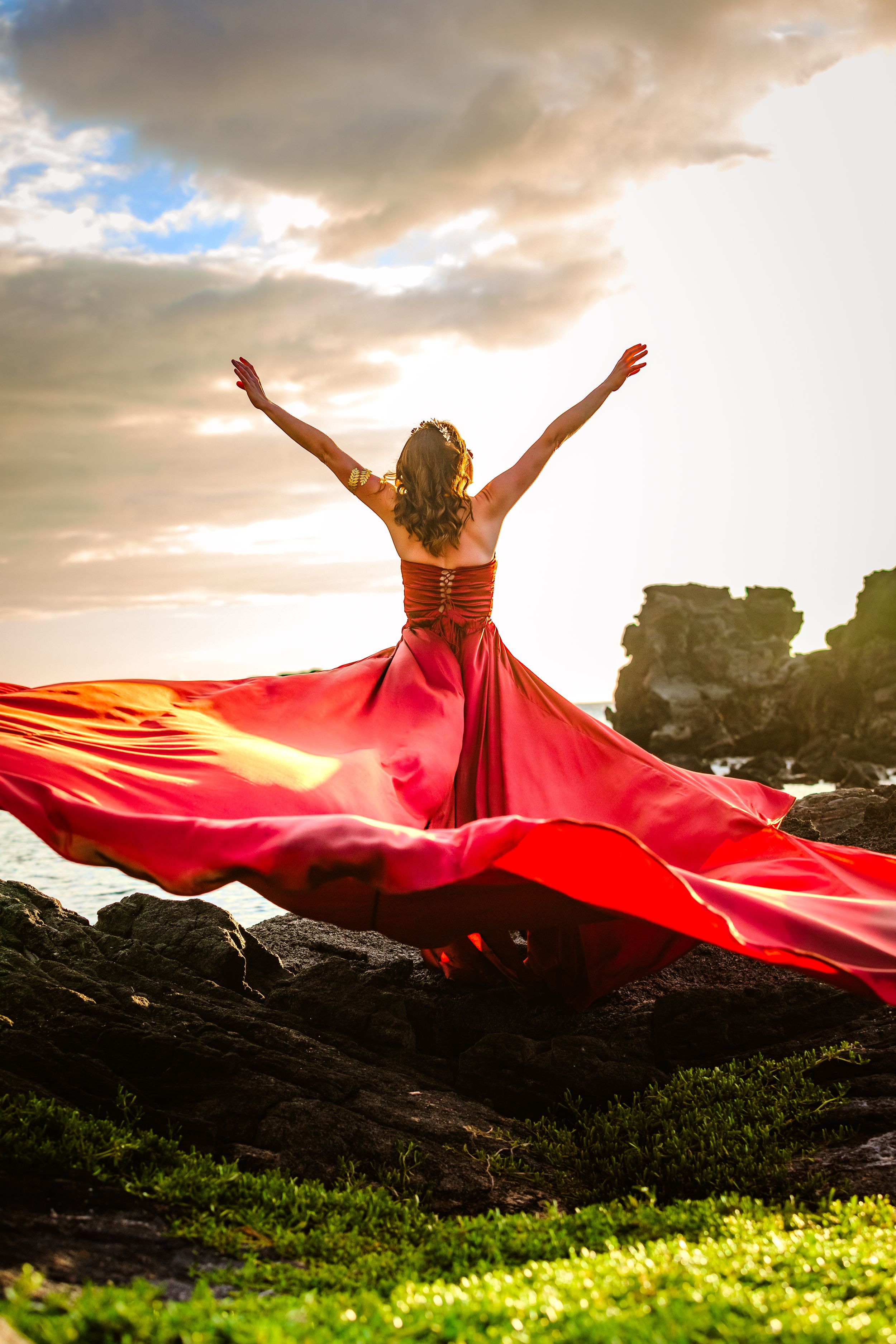 A flowing red dress catches the wind against a dramatic sunset on rocky coastal terrain.