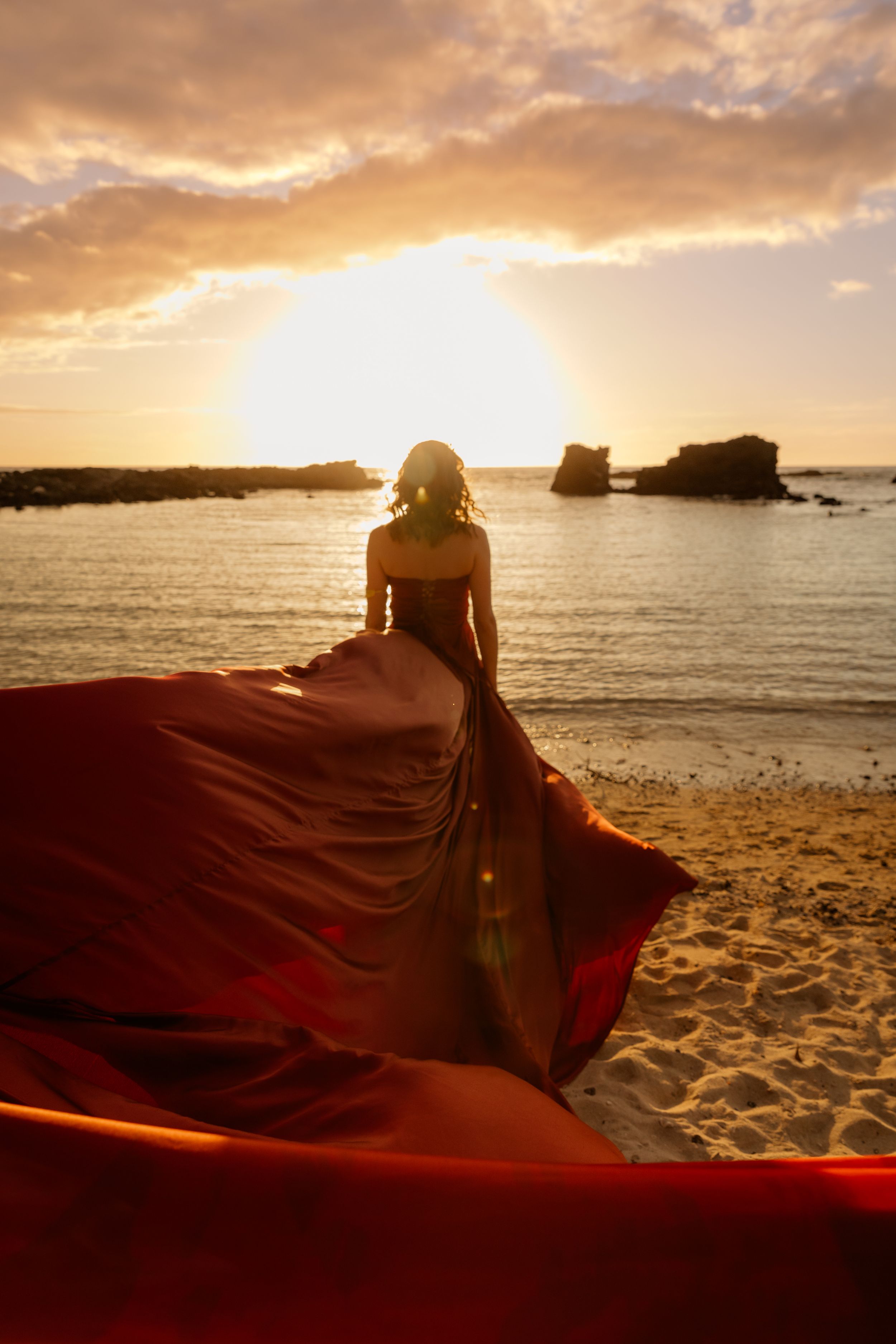 A flowing red dress silhouetted against golden sunset over beach and rock formations.