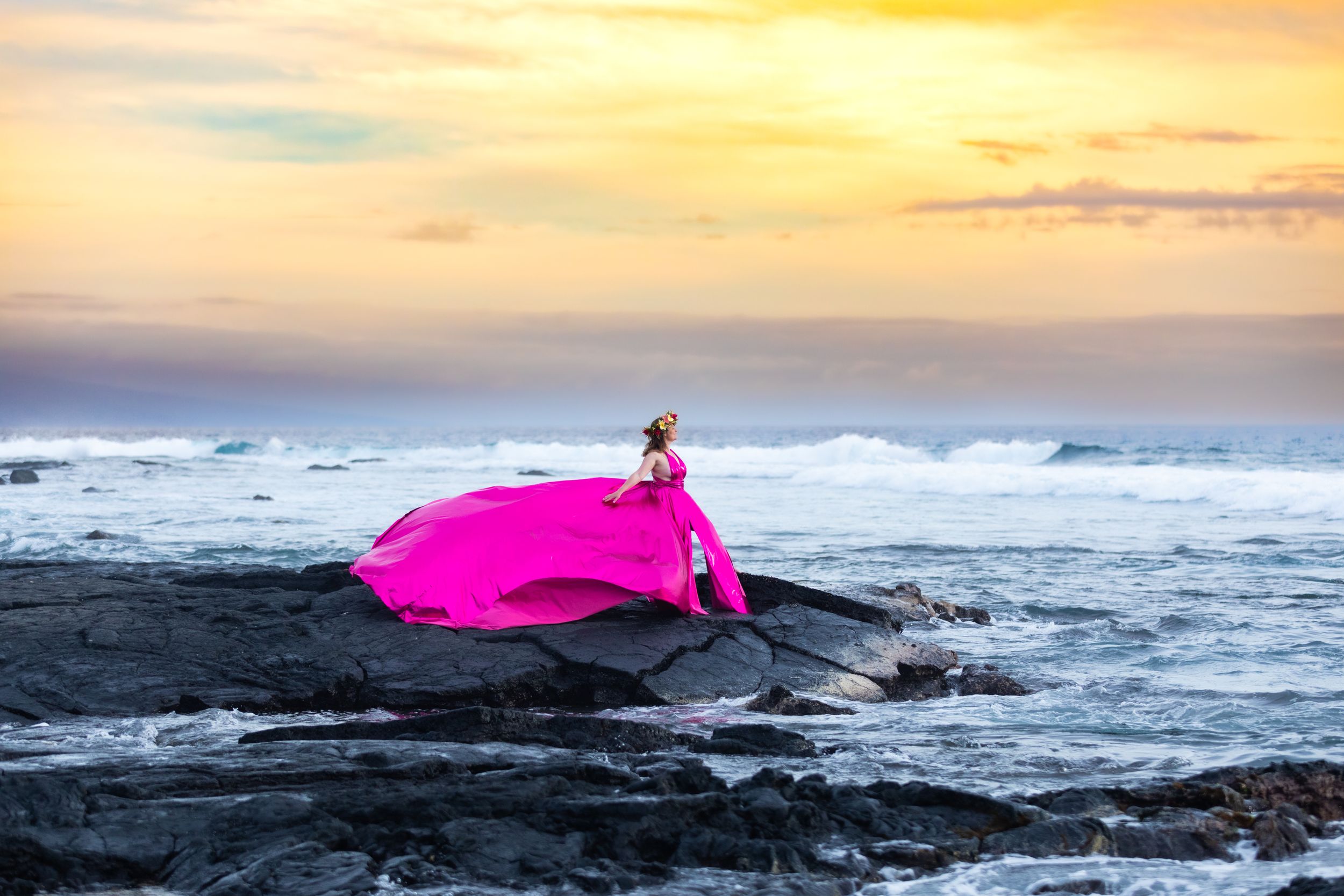 A bright pink fabric flows dramatically over black rocks at sunset along a dramatic coastline.