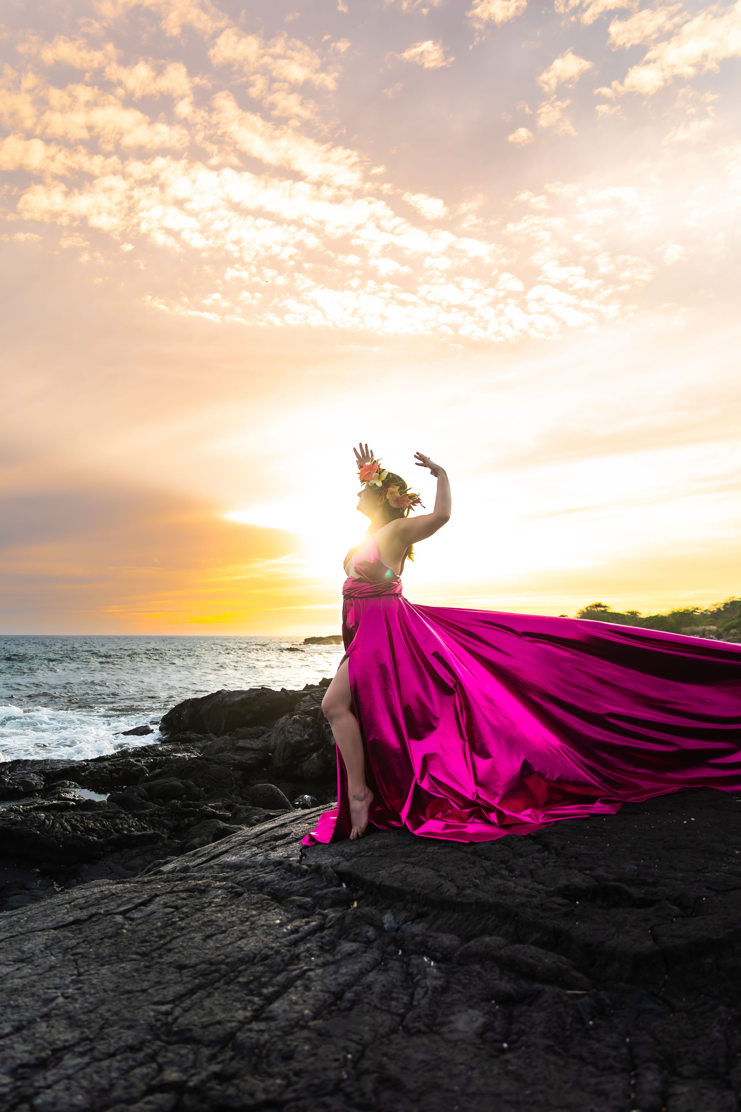 A magenta dress billows dramatically on coastal rocks during golden sunset.