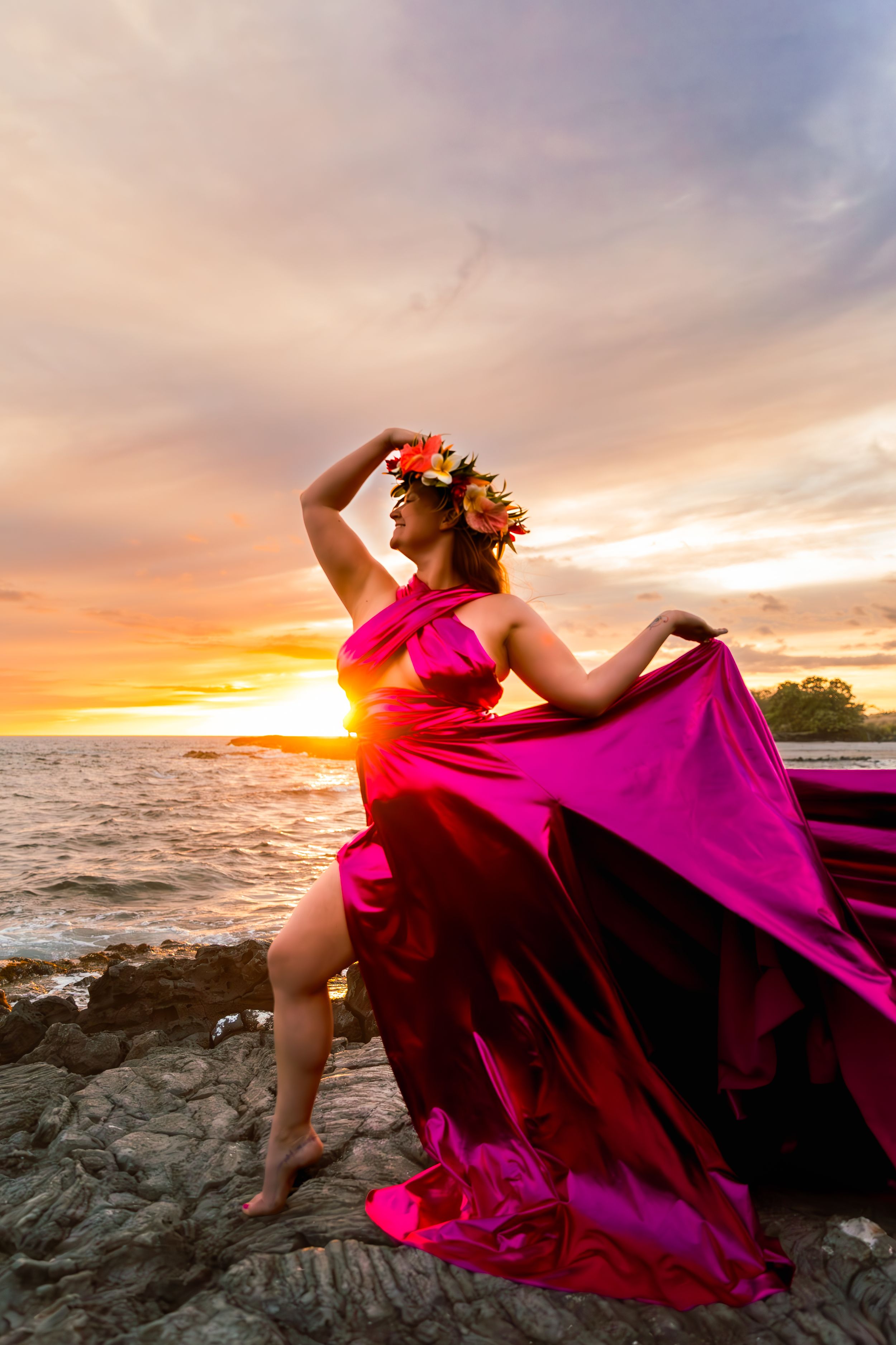 A vibrant pink dress flows in ocean breeze as sun sets over volcanic coastline.