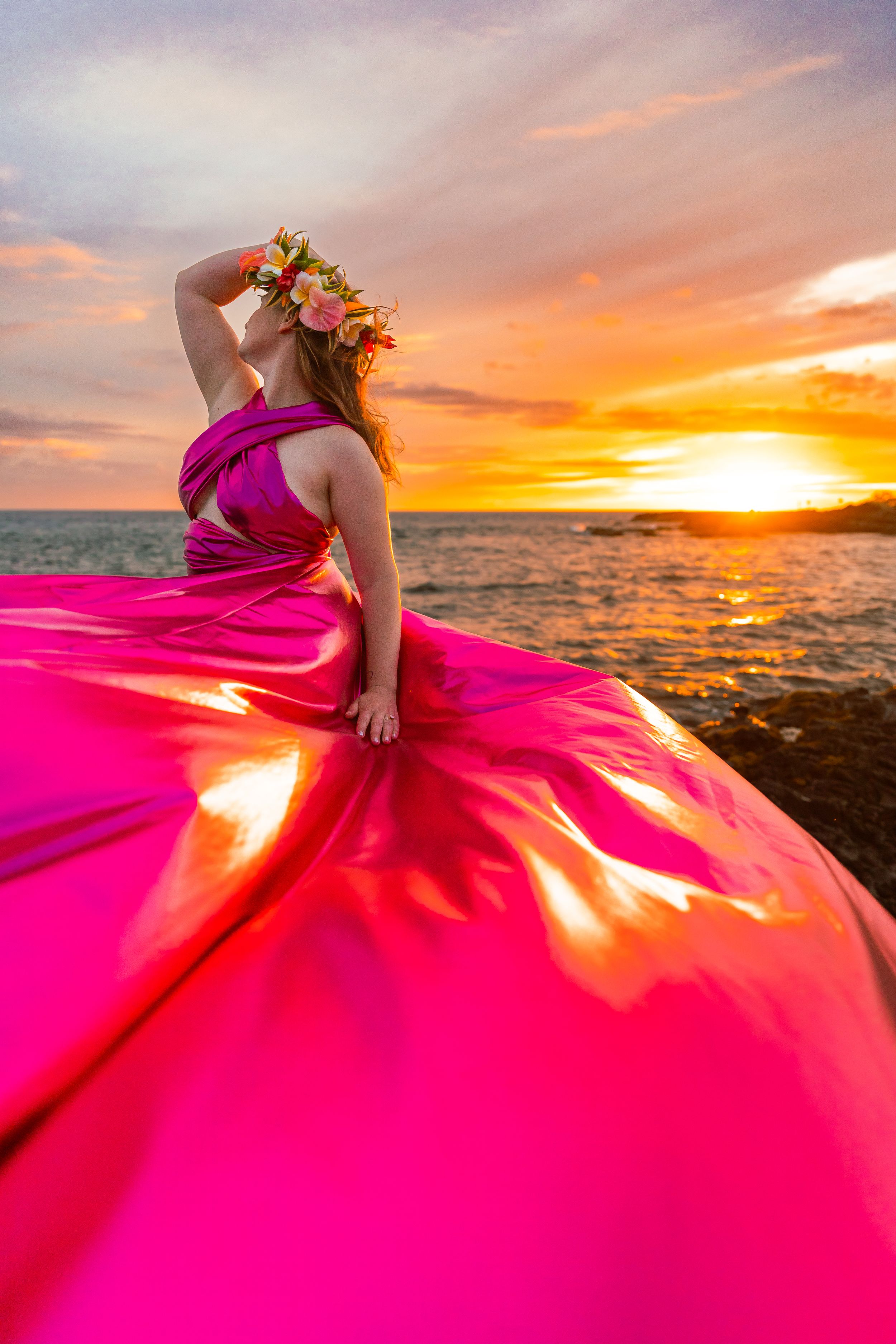 A flowing pink gown catches sunlight dramatically against vivid orange sunset over ocean.
