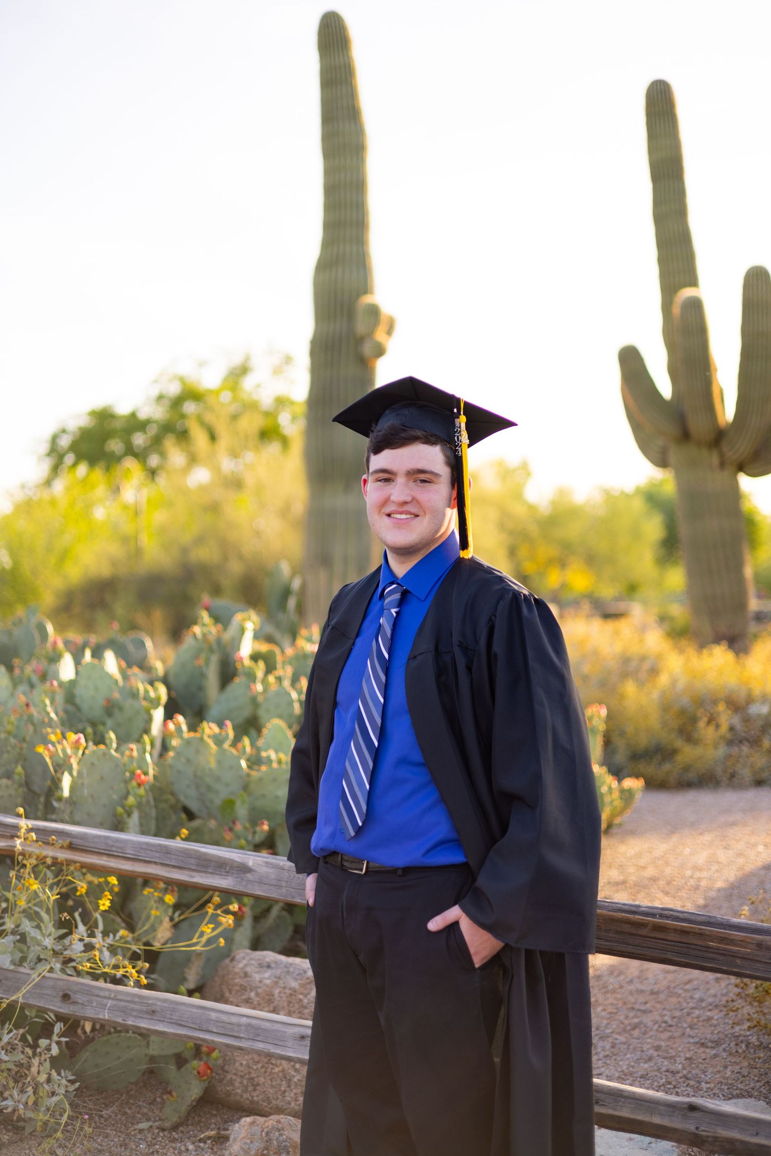 Graduate in cap and gown poses by saguaro cactus in desert landscape.