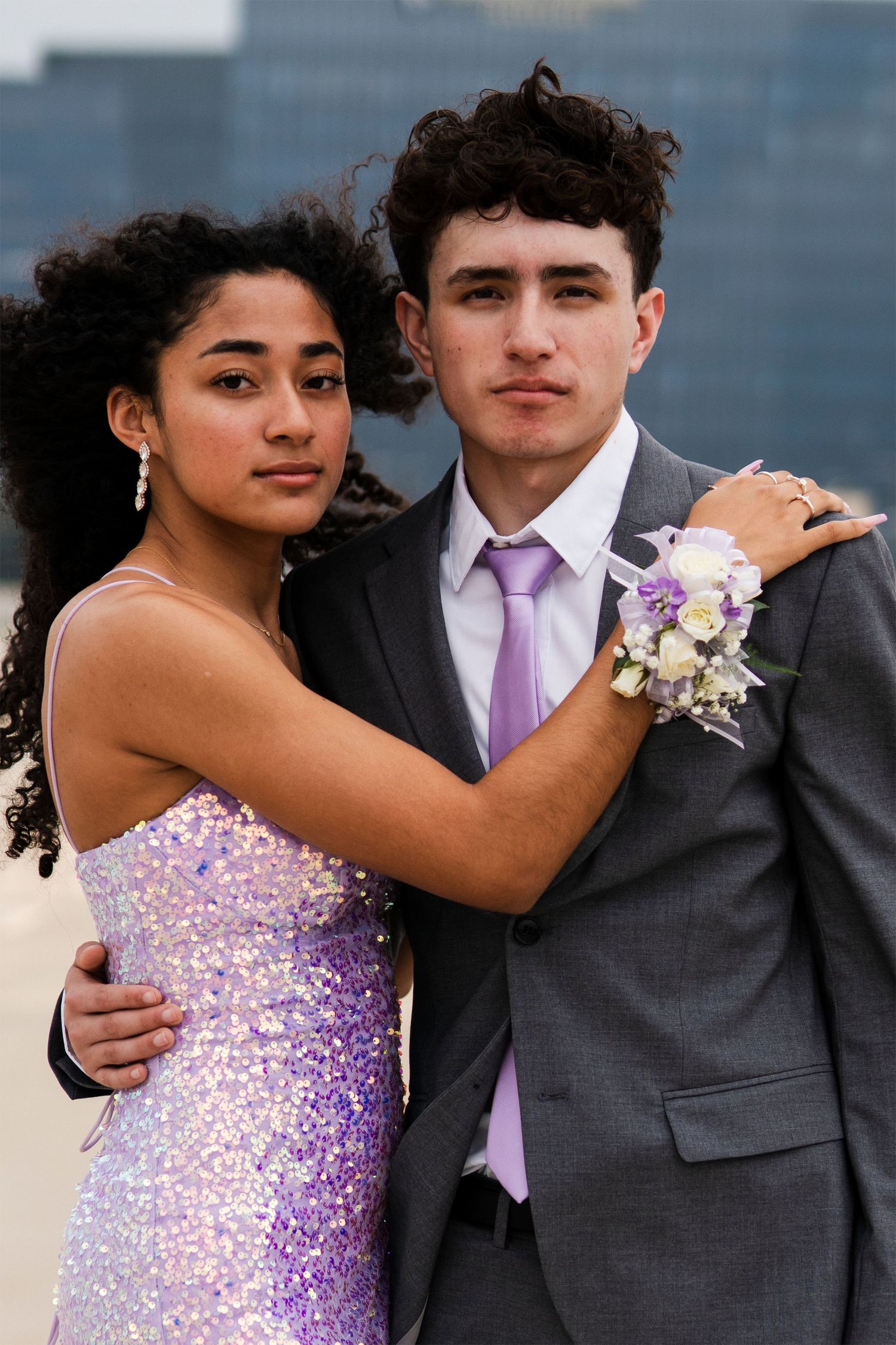 A formal couple poses in prom attire with a purple sequin dress and corsage against a city backdrop.