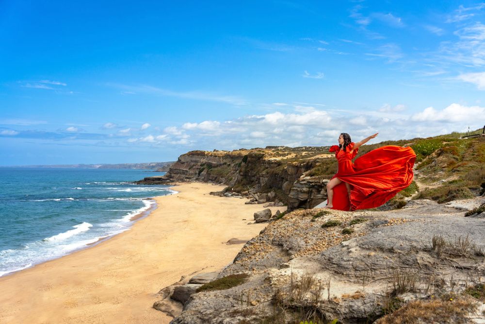 A flowing red dress billows dramatically against rocky coastal cliffs and sandy beach under blue skies.