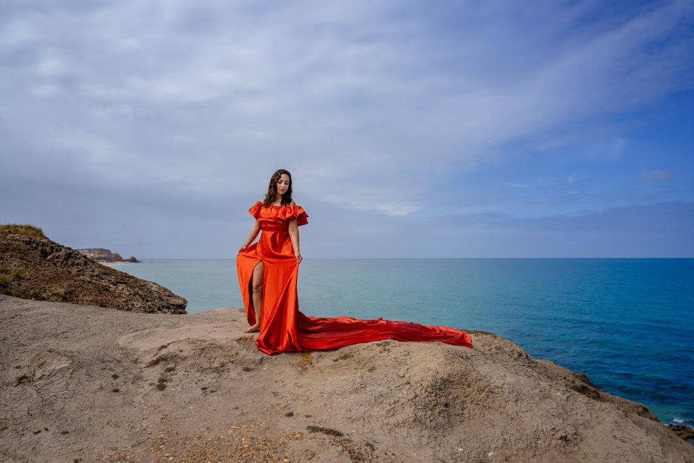 A vibrant orange gown flows in the wind on a dramatic coastal cliff overlooking turquoise waters.
