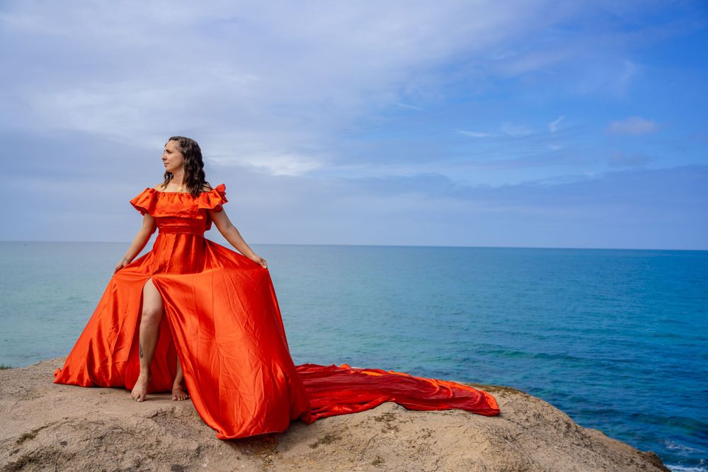 A dramatic red dress catches the wind on a rocky ocean overlook against a cloudy sky.