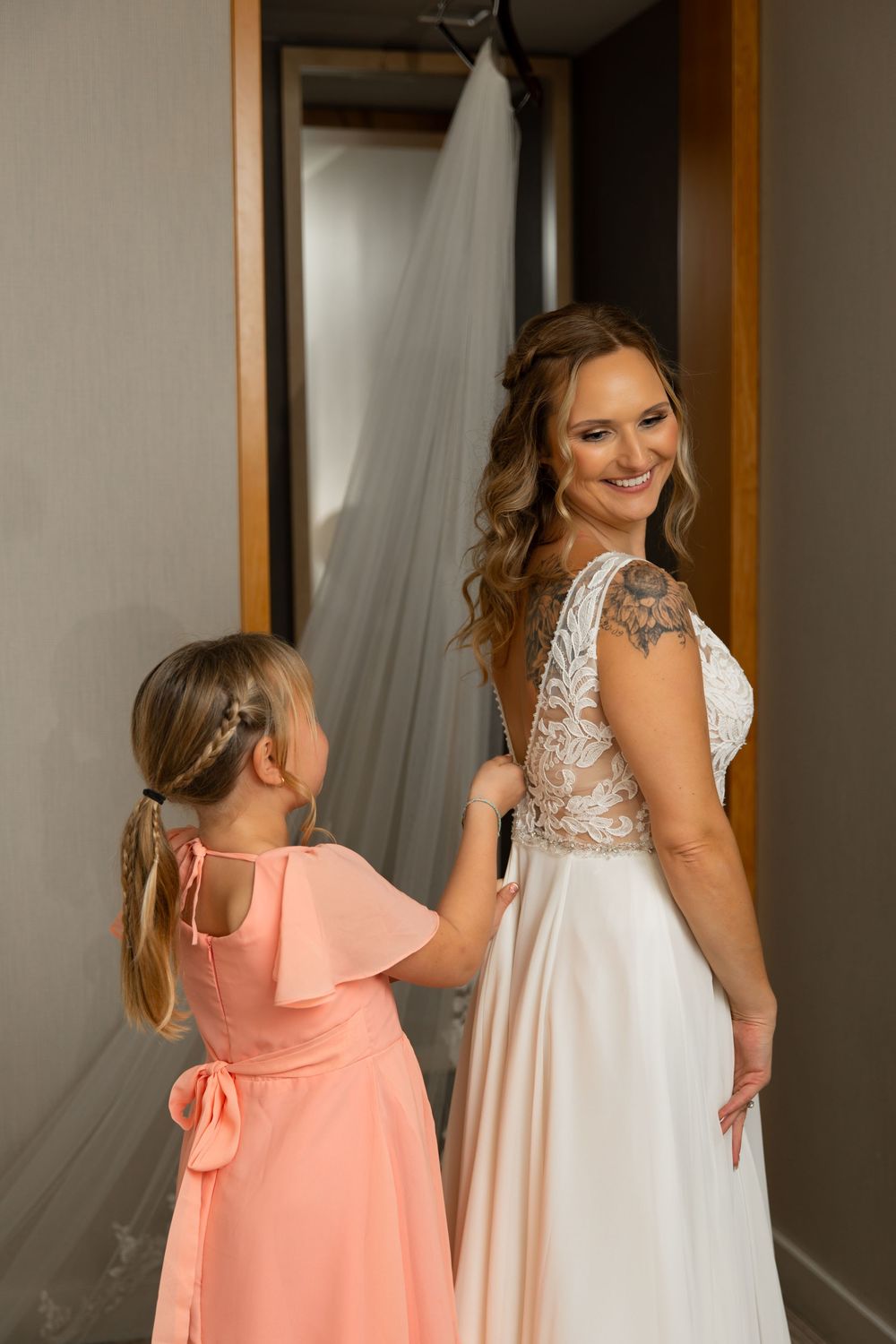 Two young girls in white and coral dresses twirl and play together in a room with sheer curtains.