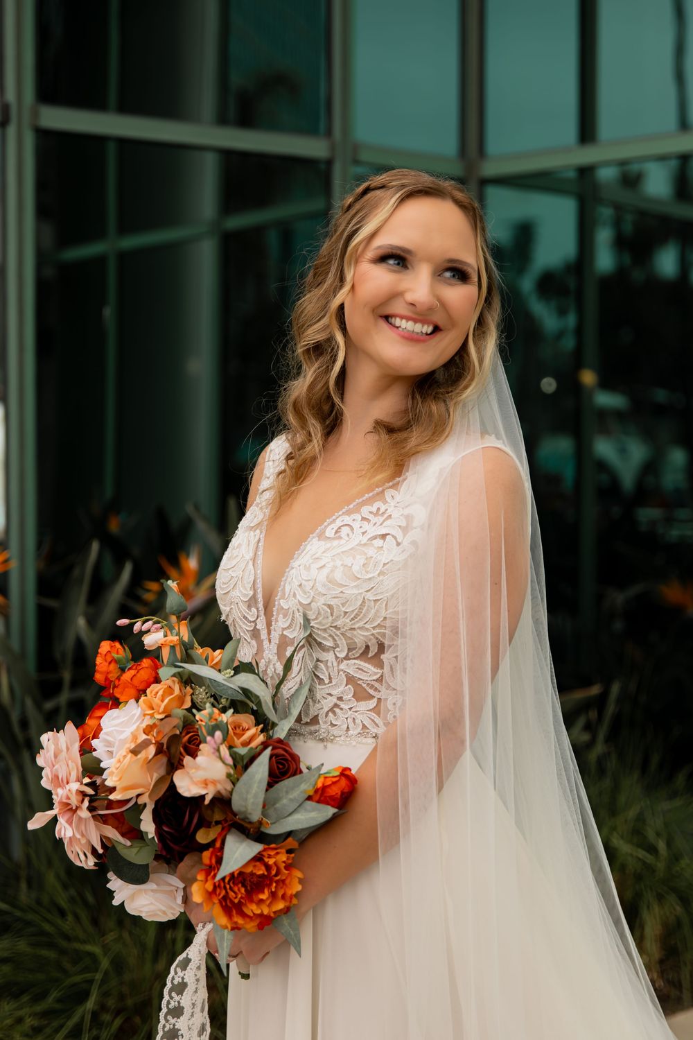 Bride in lace wedding dress with plunging neckline holds vibrant orange and pink floral bouquet against modern building.