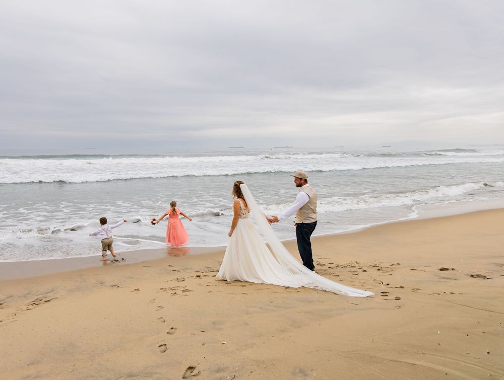 A couple celebrates their wedding day on a cloudy beach with waves crashing in the background as others walk nearby.