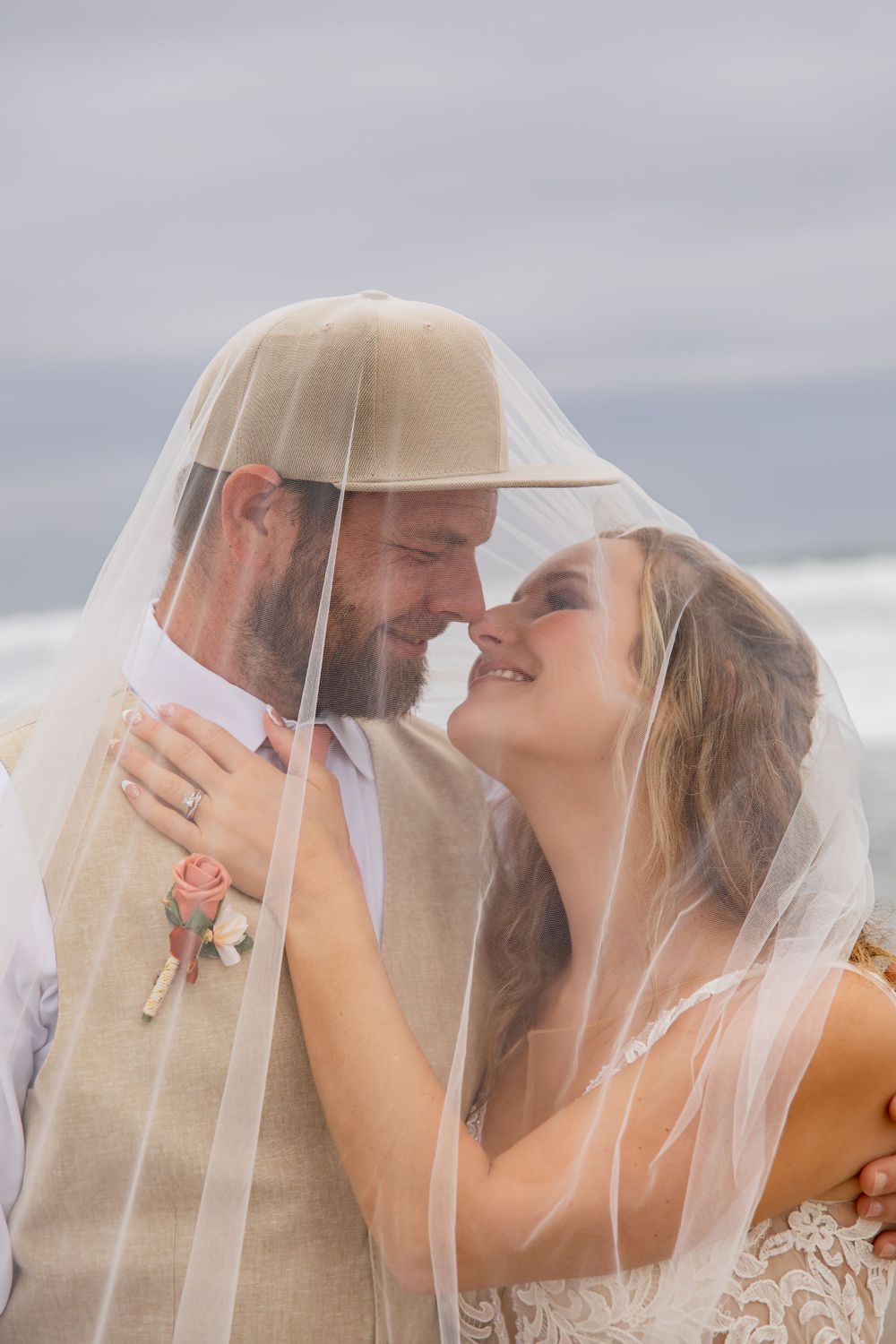 A tender moment shared under a flowing veil against an overcast beach backdrop.