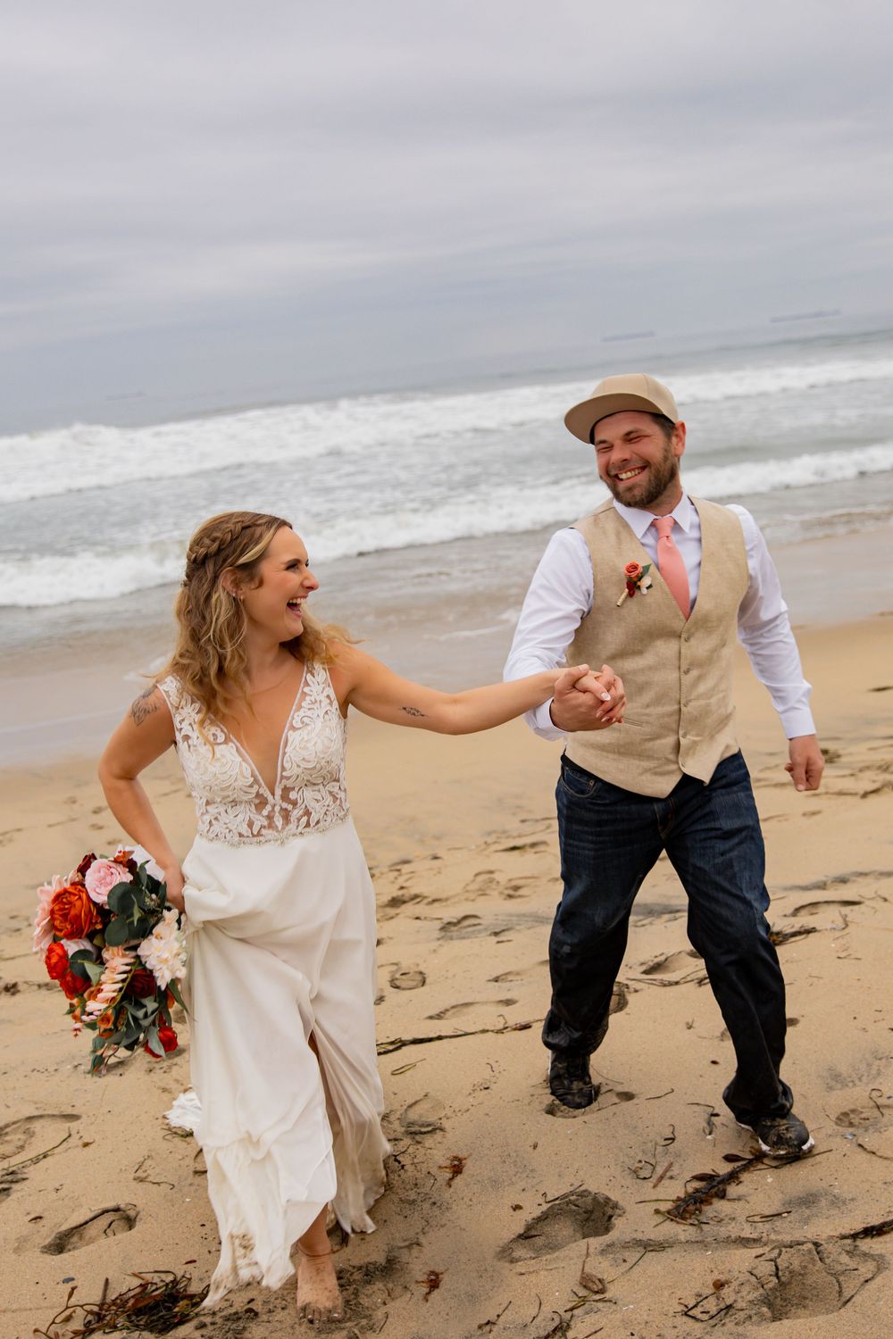 A joyful couple in wedding attire dances and laughs together on a cloudy beach with crashing waves in the background.