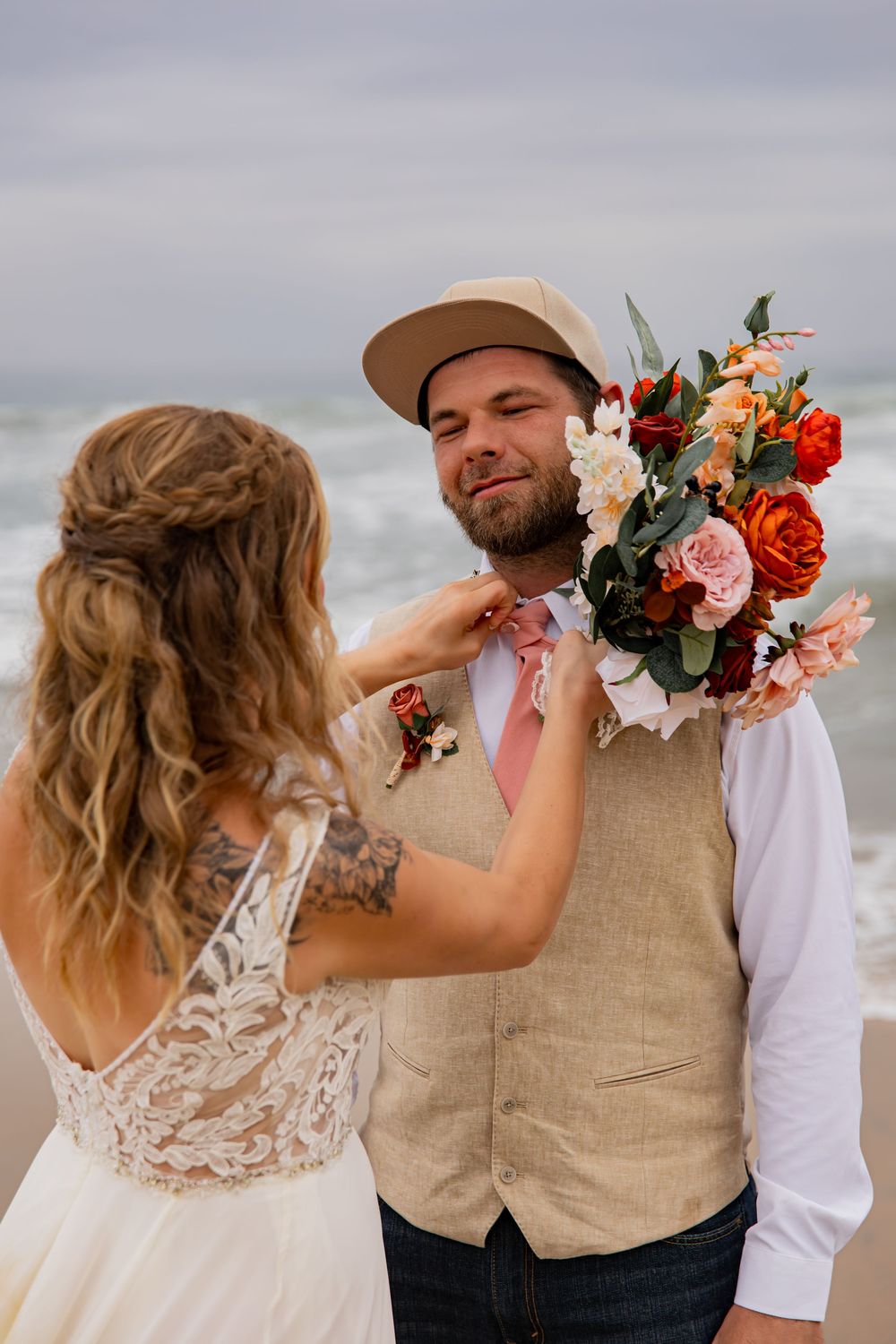 A couple shares playful moments on their wedding day wearing a lace gown and tan vest on a cloudy beach.