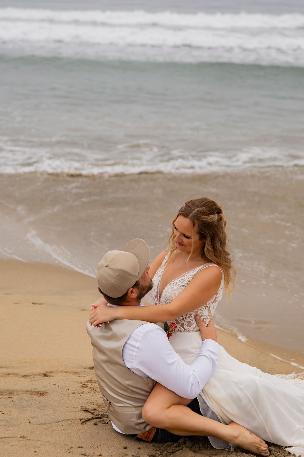 A couple in wedding attire playfully poses together on a sandy beach with ocean waves in the background.