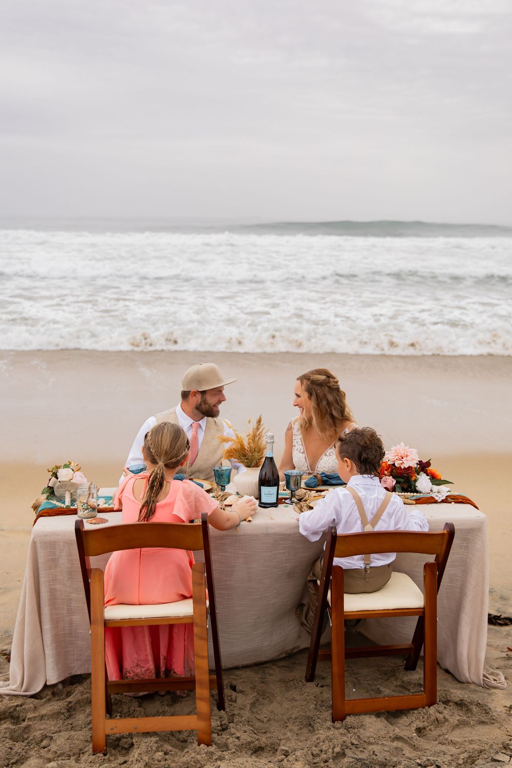 A romantic beach dinner setup with wooden chairs and tables draped in neutral linens along the sandy shoreline.