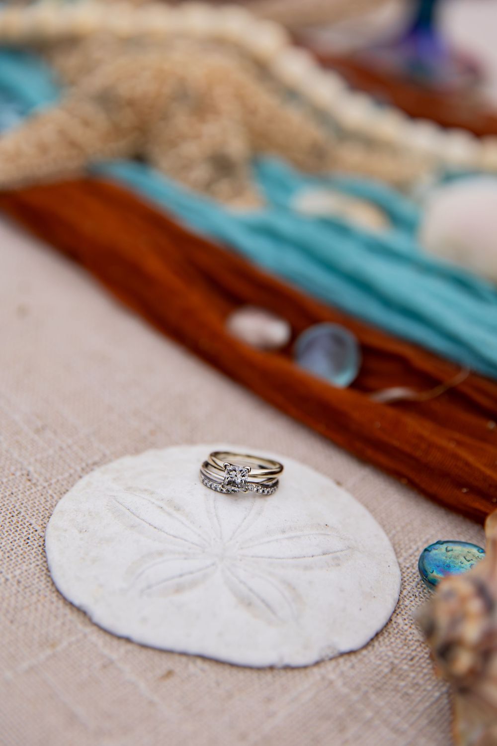A wedding ring sits atop a white sand dollar on a rustic wooden surface with turquoise accents.