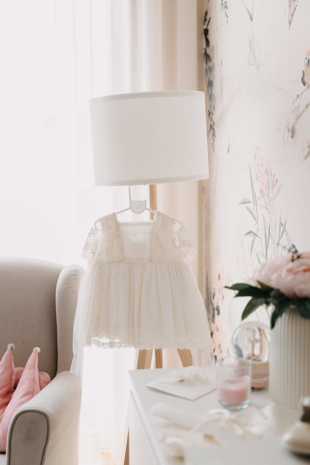White christening dress displayed near table lamp with soft pink decor elements.