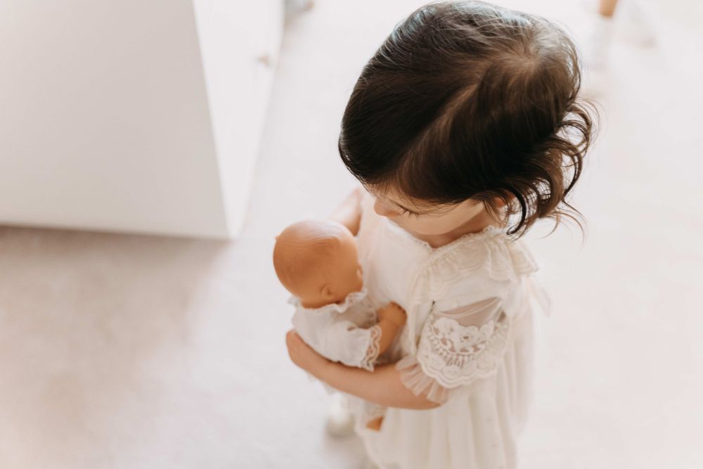 A small child in white dress holding a baby doll while standing in a bright room.