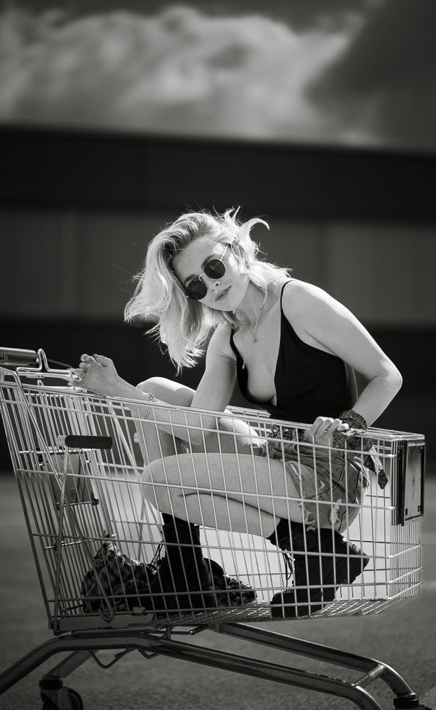 Black and white image of a blonde woman sitting inside a shopping cart wearing sunglasses and a black bodysuit.