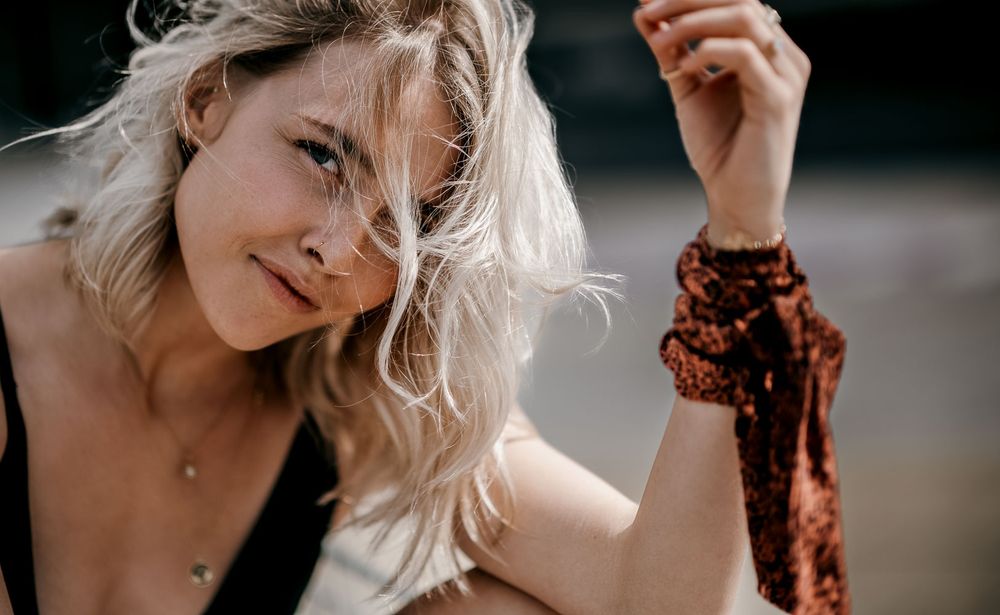 Close-up portrait of a blonde woman in a black top with a rust scarf on her wrist smiling softly in natural light.