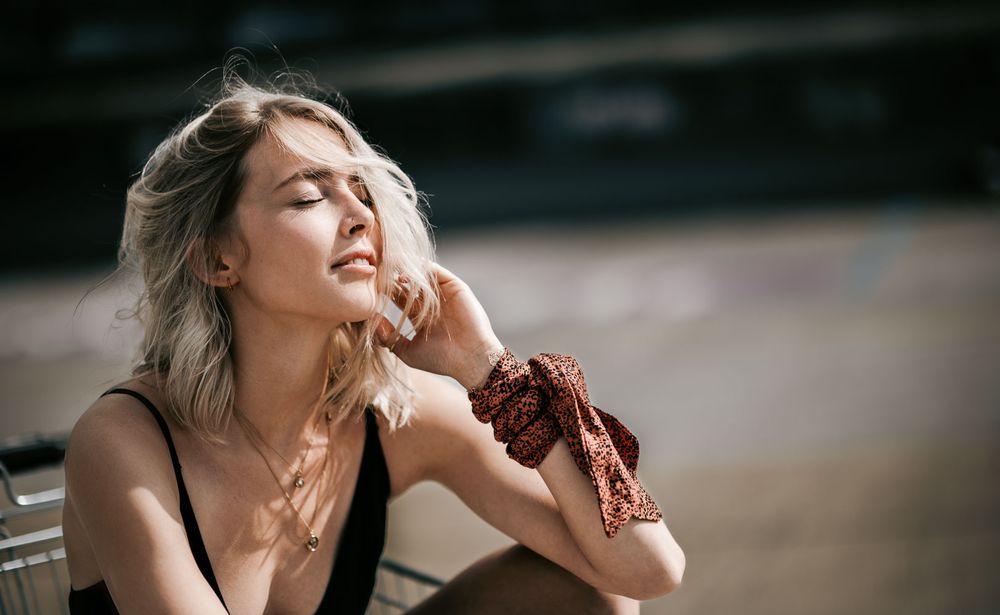 Blonde woman in a black top with a rust-colored scarf on her wrist enjoying warm sunlight with eyes closed outdoors.