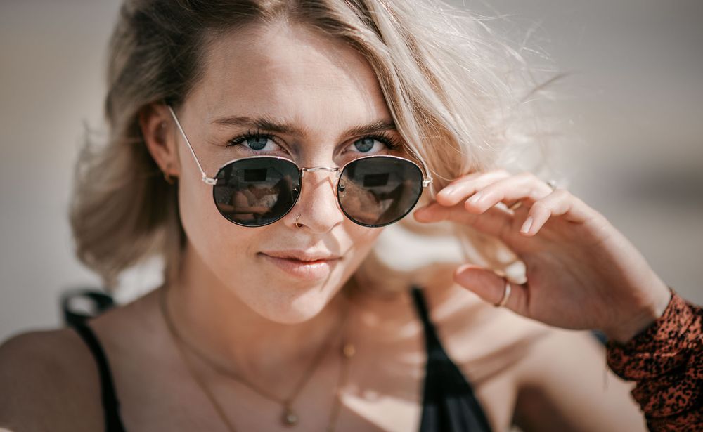 Close-up portrait of a young blonde woman peering over round sunglasses with a rust-colored scarf on her wrist.