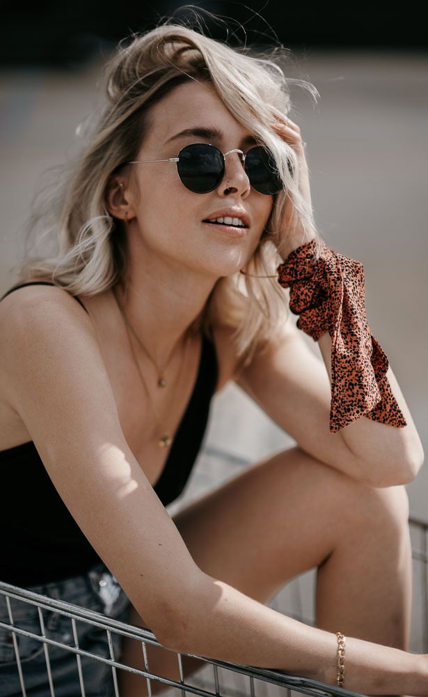 Blonde woman in black bodysuit and leopard scrunchie wearing round sunglasses outdoors.