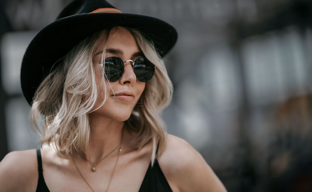 Stylish blonde woman in black hat and round sunglasses wearing a black top.