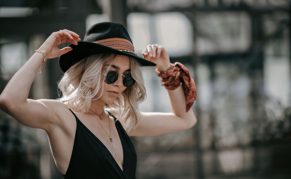 Blonde woman adjusting black wide-brim hat with leopard scarf and round sunglasses.