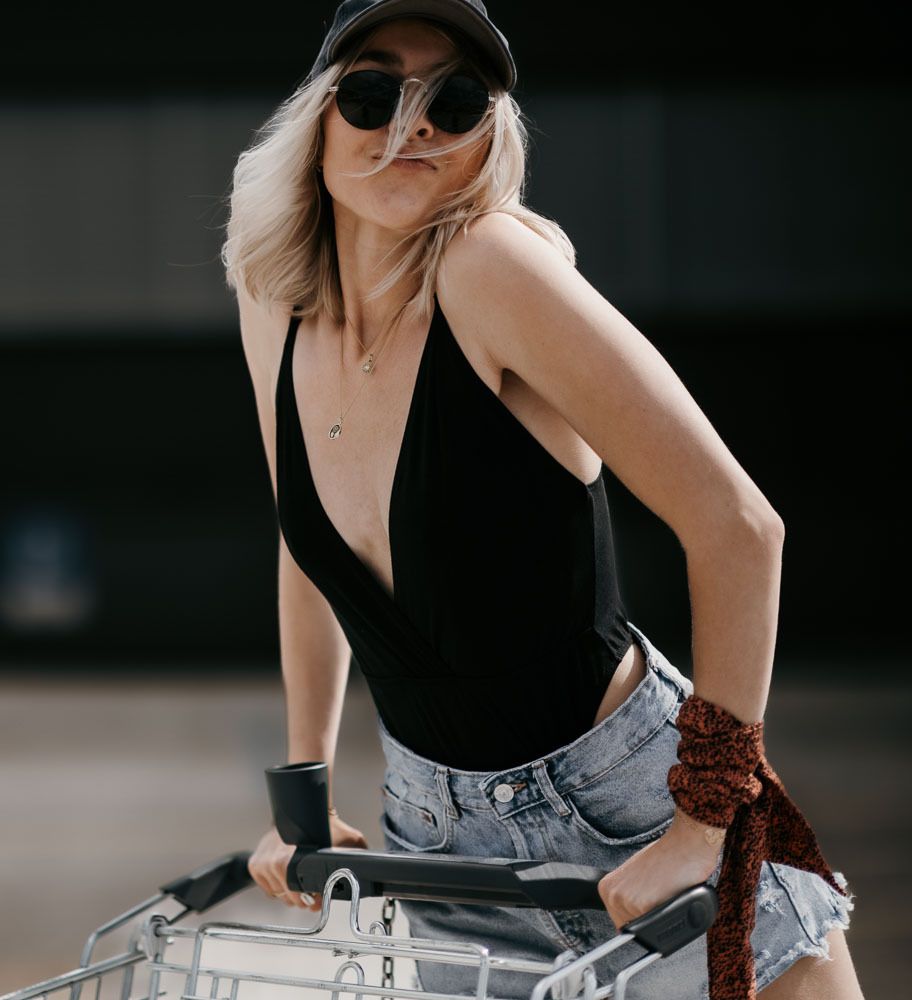 Stylish blonde woman in black bodysuit and denim shorts posing with a shopping cart wearing sunglasses and a black cap.