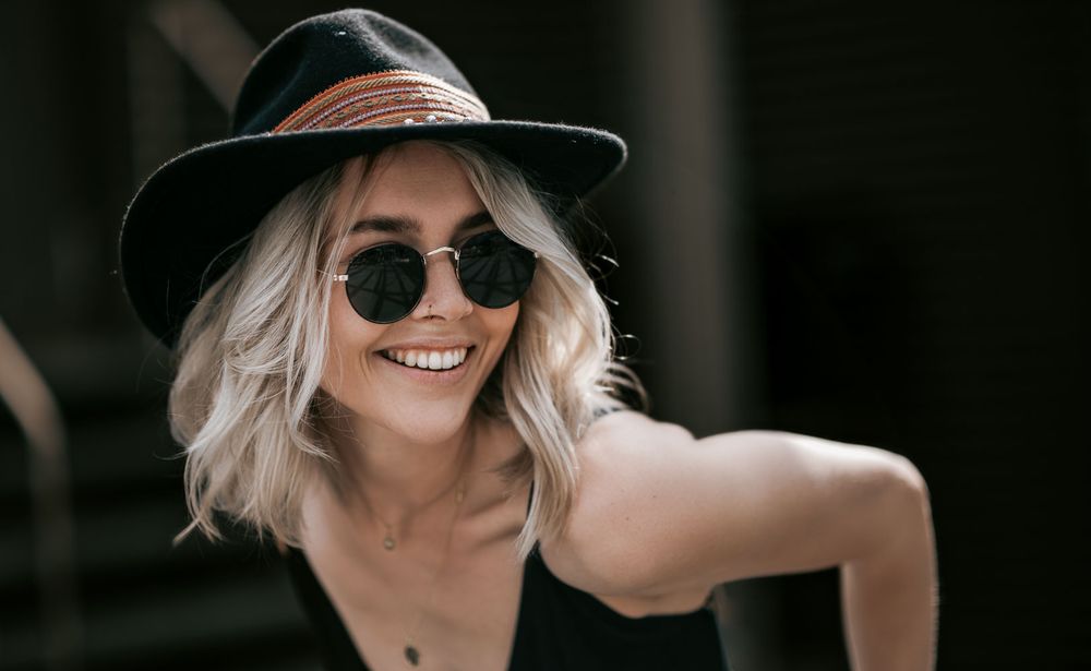 Cheerful blonde woman smiling in black fedora and round sunglasses wearing black top.