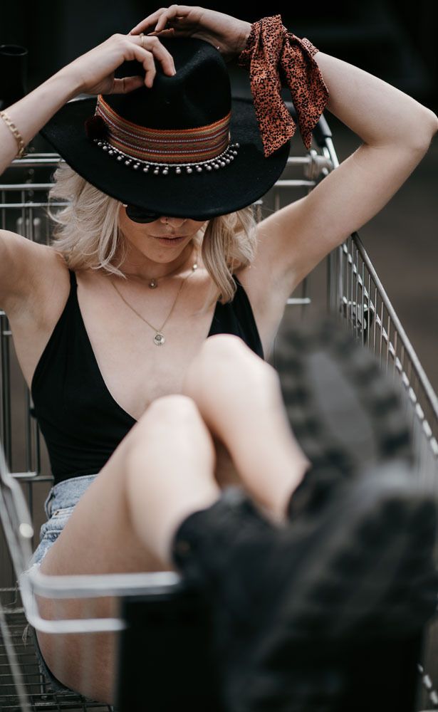Blonde woman sitting in shopping cart wearing black hat with leopard scarf and bodysuit.