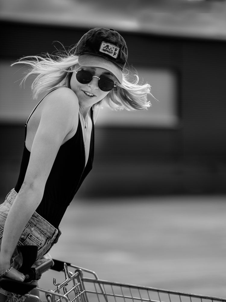 Black and white photo of a blonde woman in sunglasses and cap leaning on a shopping cart with hair blowing in the wind.