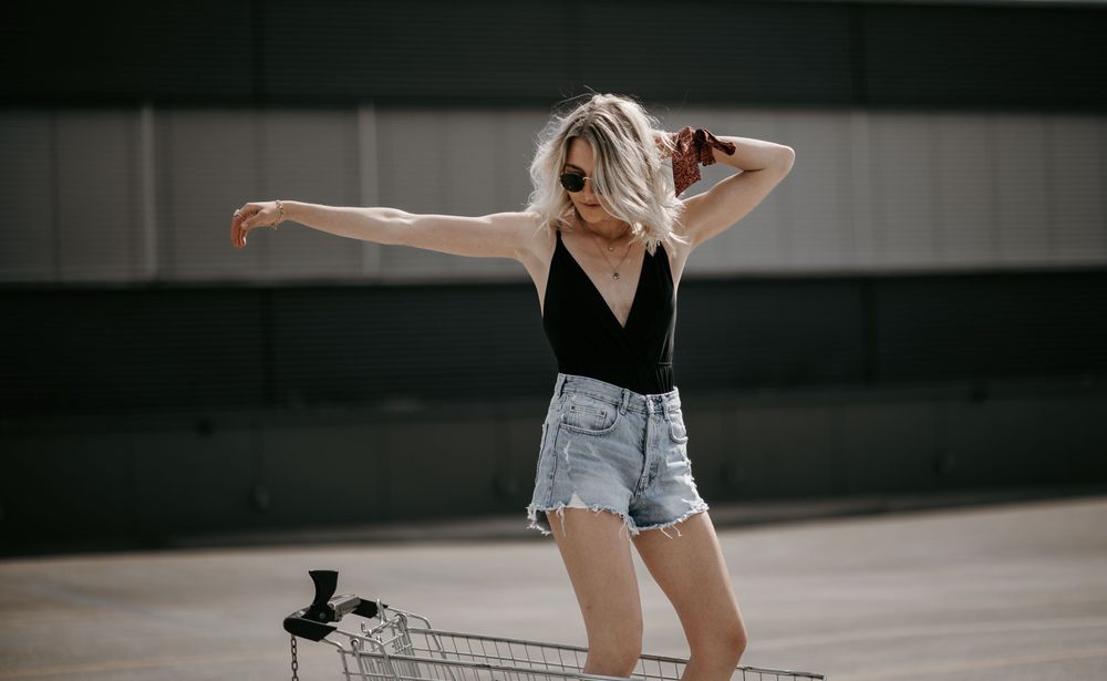 Blonde woman in black bodysuit and denim shorts posing outdoors near a shopping cart with arms outstretched in sunlight.