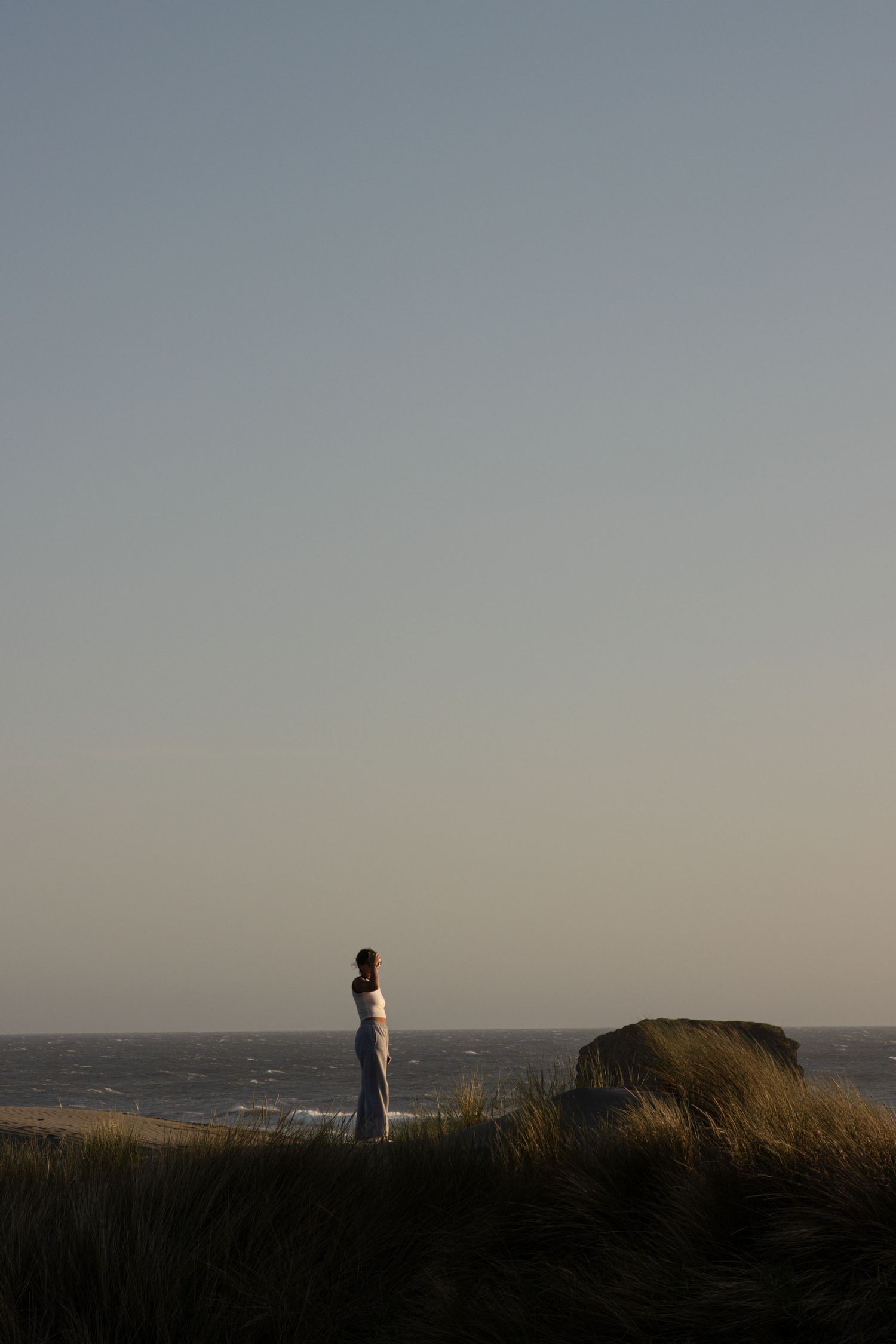 Cora Emilie Media Solitary figure stands on coastal cliff edge against dusky sky at sunset.
