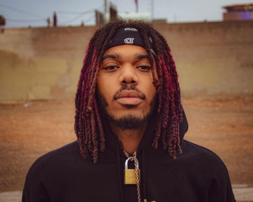 Portrait of a person with purple dreadlocks wearing a black hoodie and chain necklace.