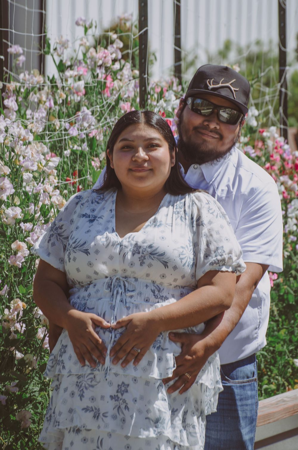 Pregnant couple poses together in a flower garden, woman in floral dress with partner in cap and sunglasses.