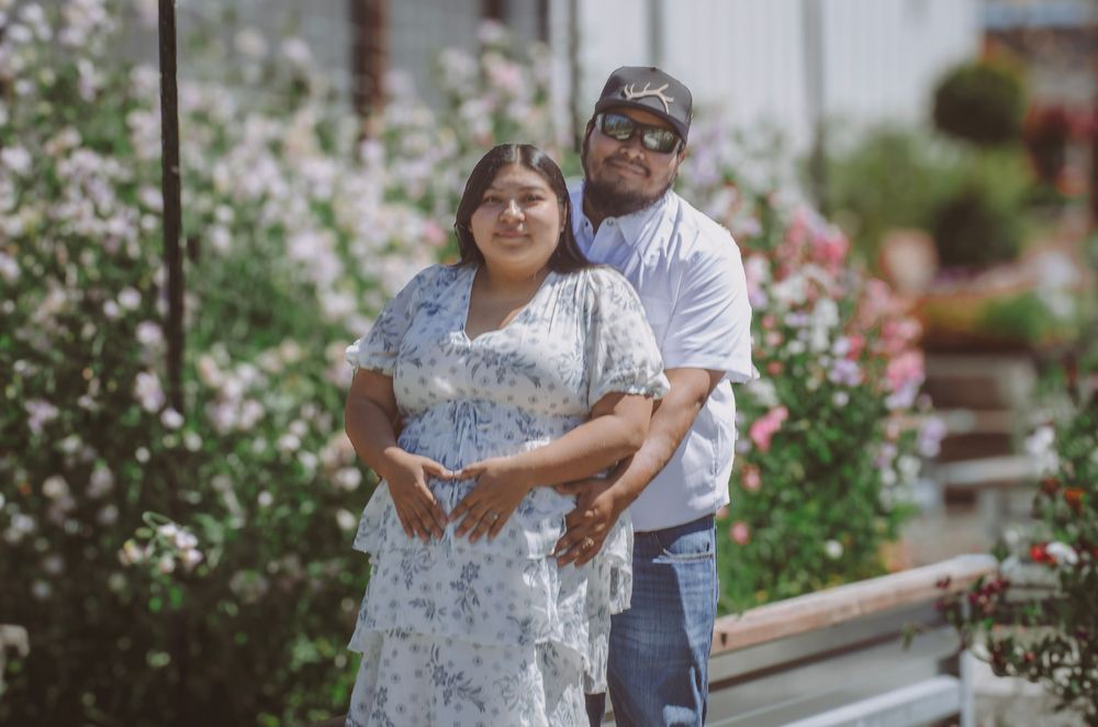 Couple posing together in front of blooming flowers, woman in floral dress, man in white shirt and cap.
