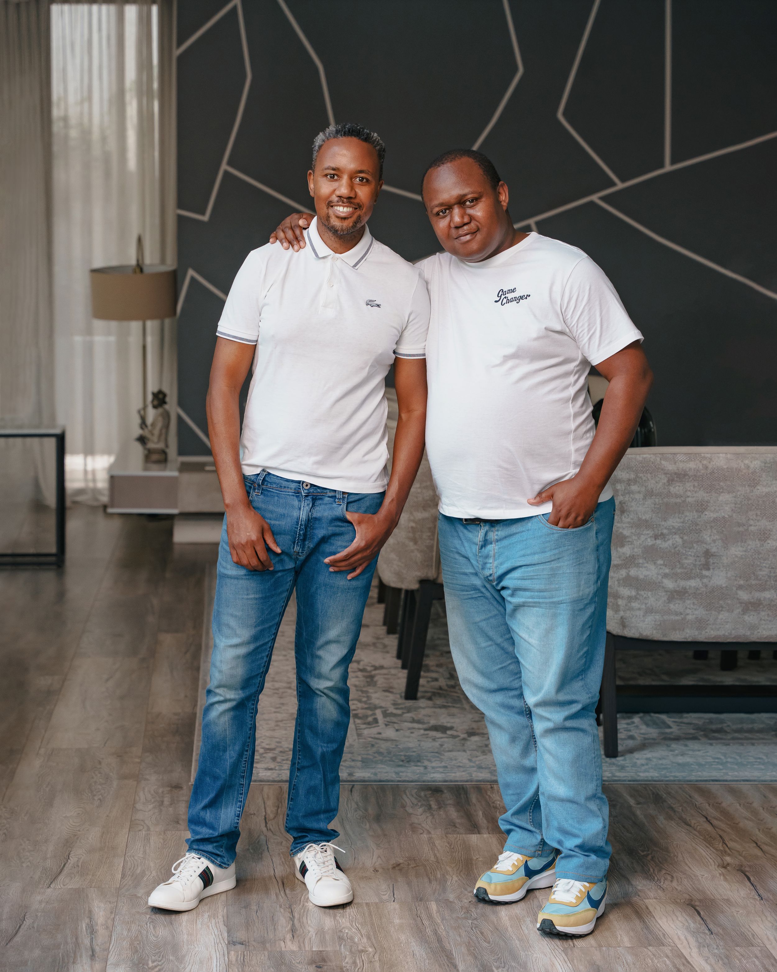 Two men in casual attire pose together against a dark wall with geometric patterns.