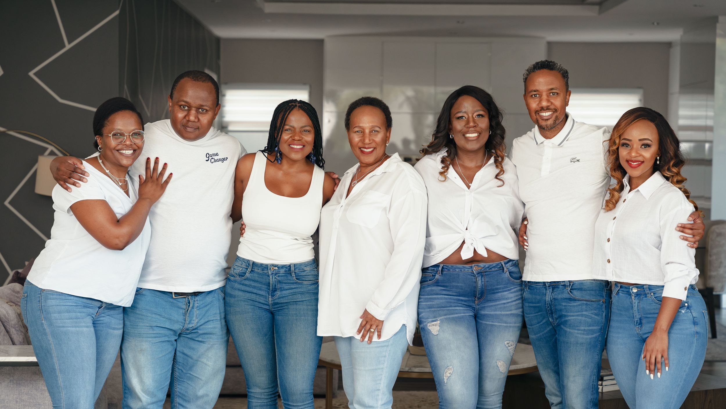 A group of people in coordinated white tops and jeans pose together against a modern wall design.