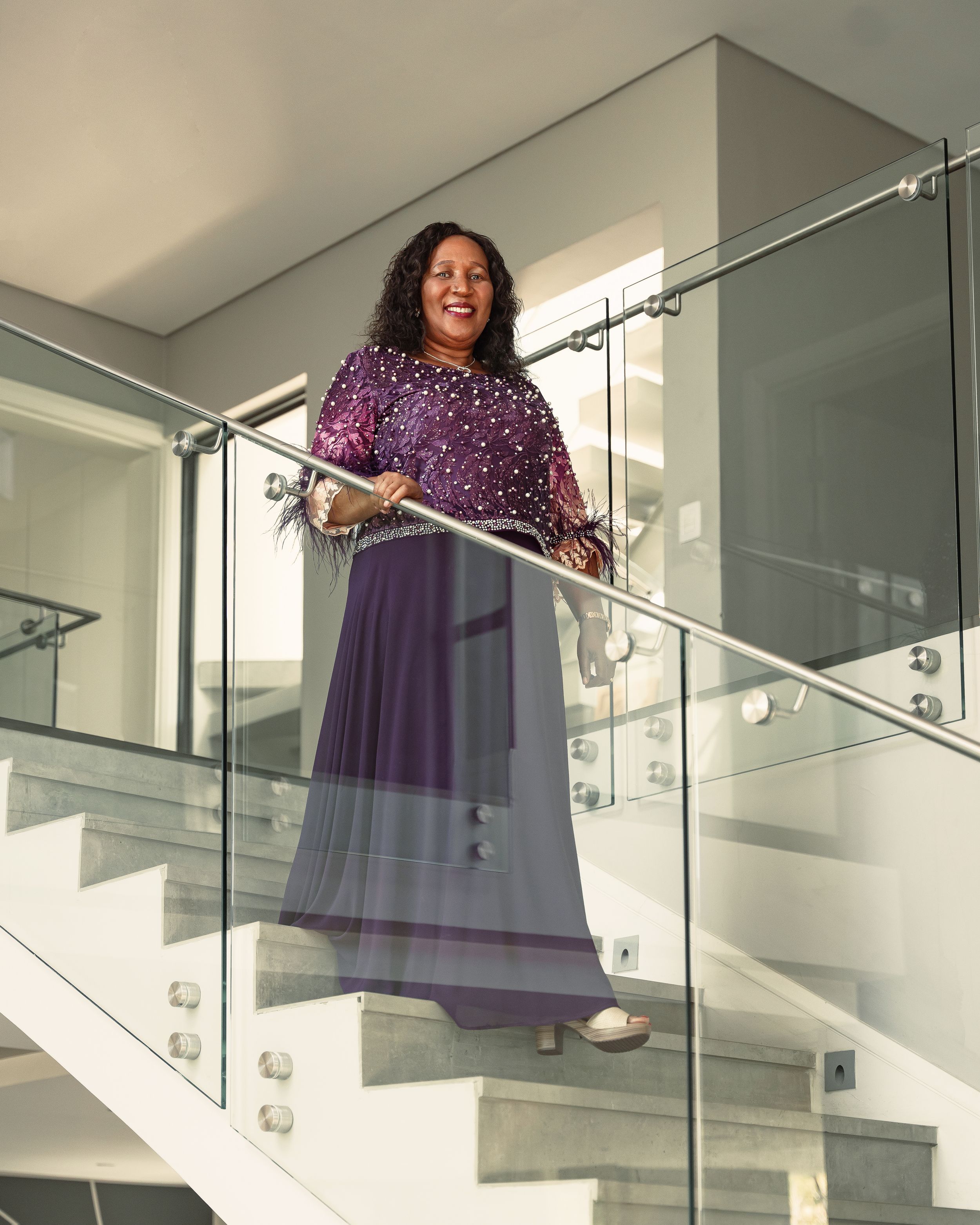 A person in a purple sequined dress poses elegantly on modern glass-railed stairs.