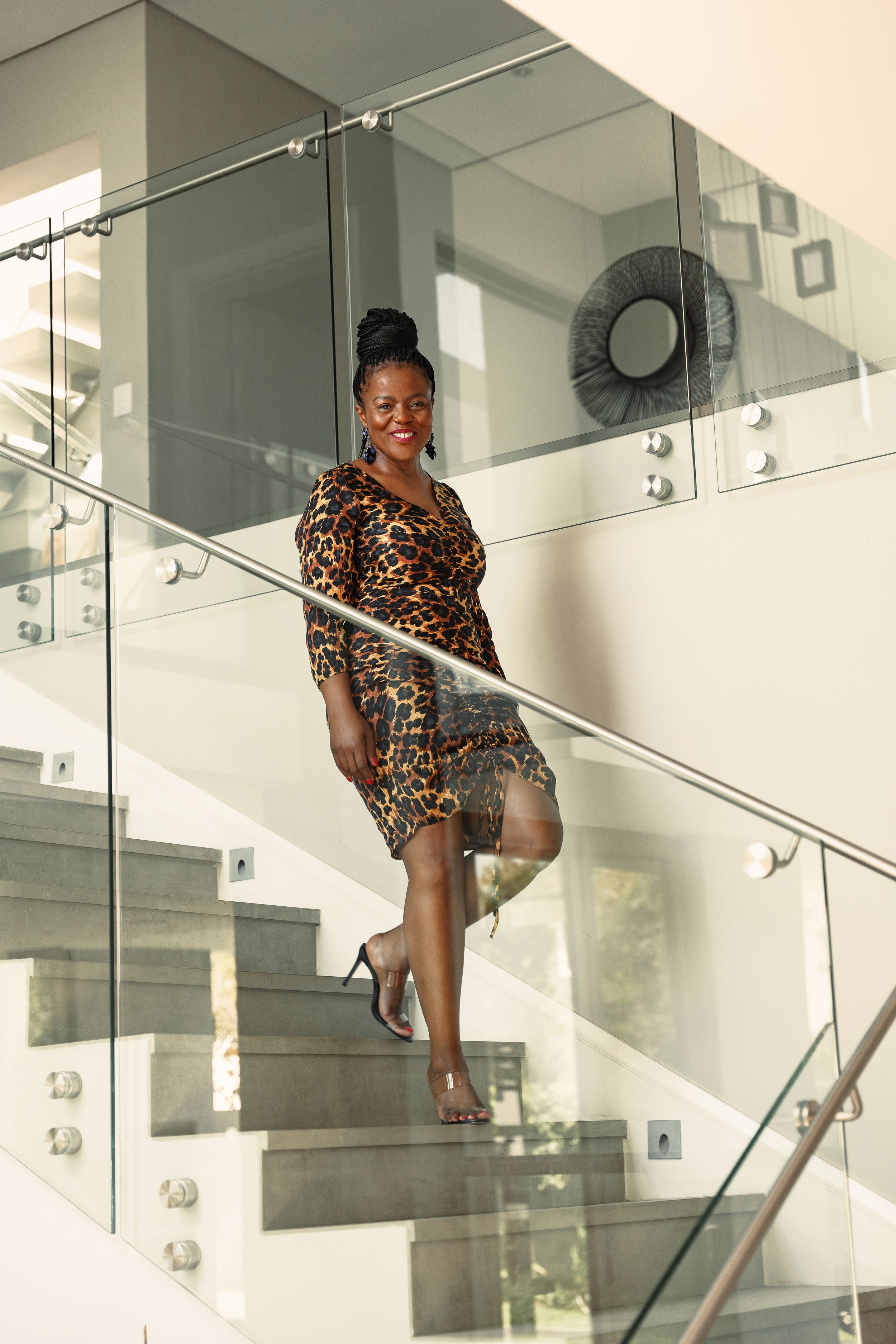 A person in an animal print dress poses confidently on a modern staircase with glass panels.