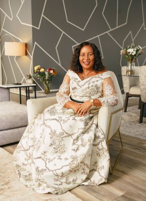 A person wearing an elegant white floral lace gown sits in a chair against a modern geometric wall design.